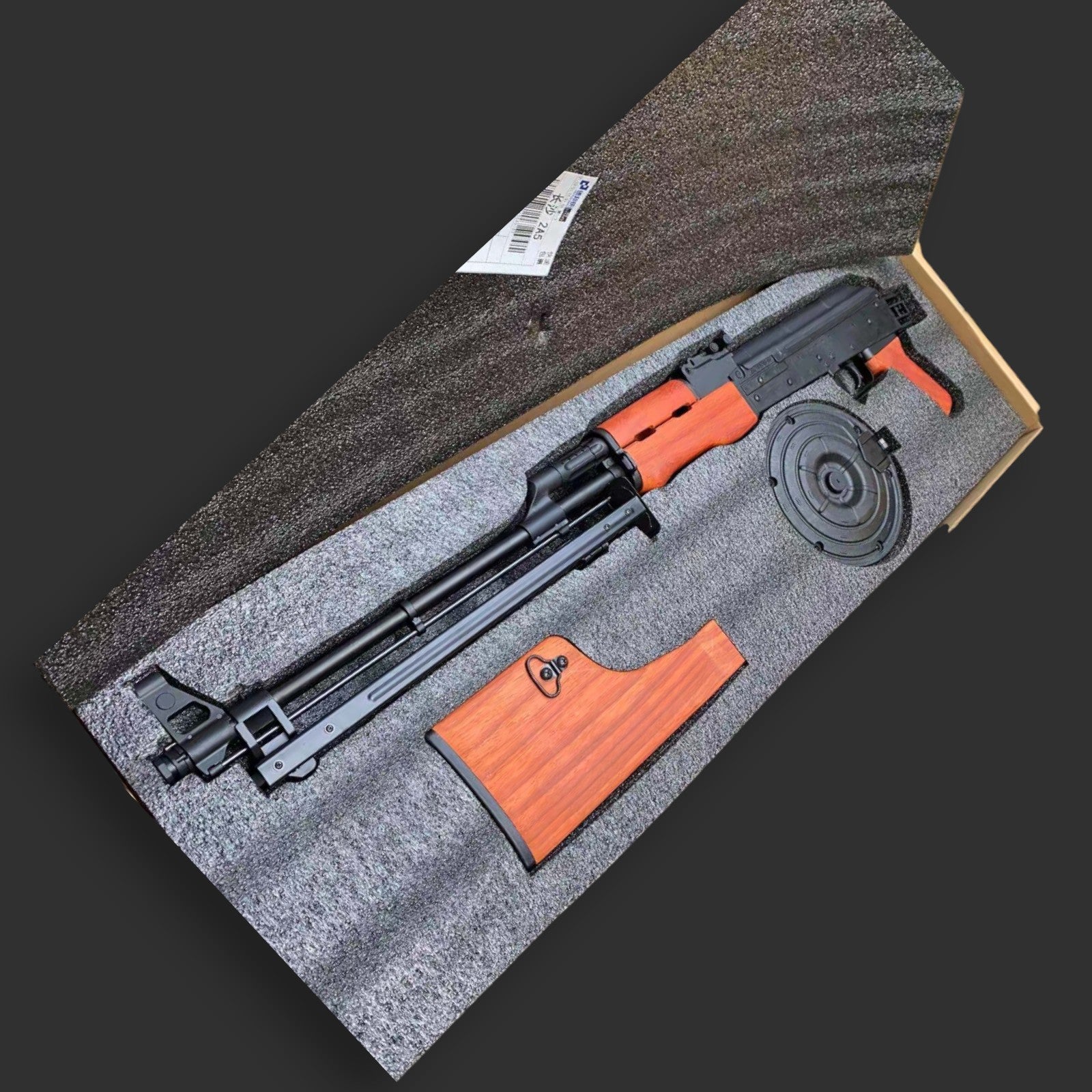 A disassembled MST RPK Gel blaster with a wooden stock and drum magazine, showcasing an authentic RPK design by BlasterMasters, is placed in a padded carrying case. A small object is seen tucked in a pocket inside the lid of the case.
