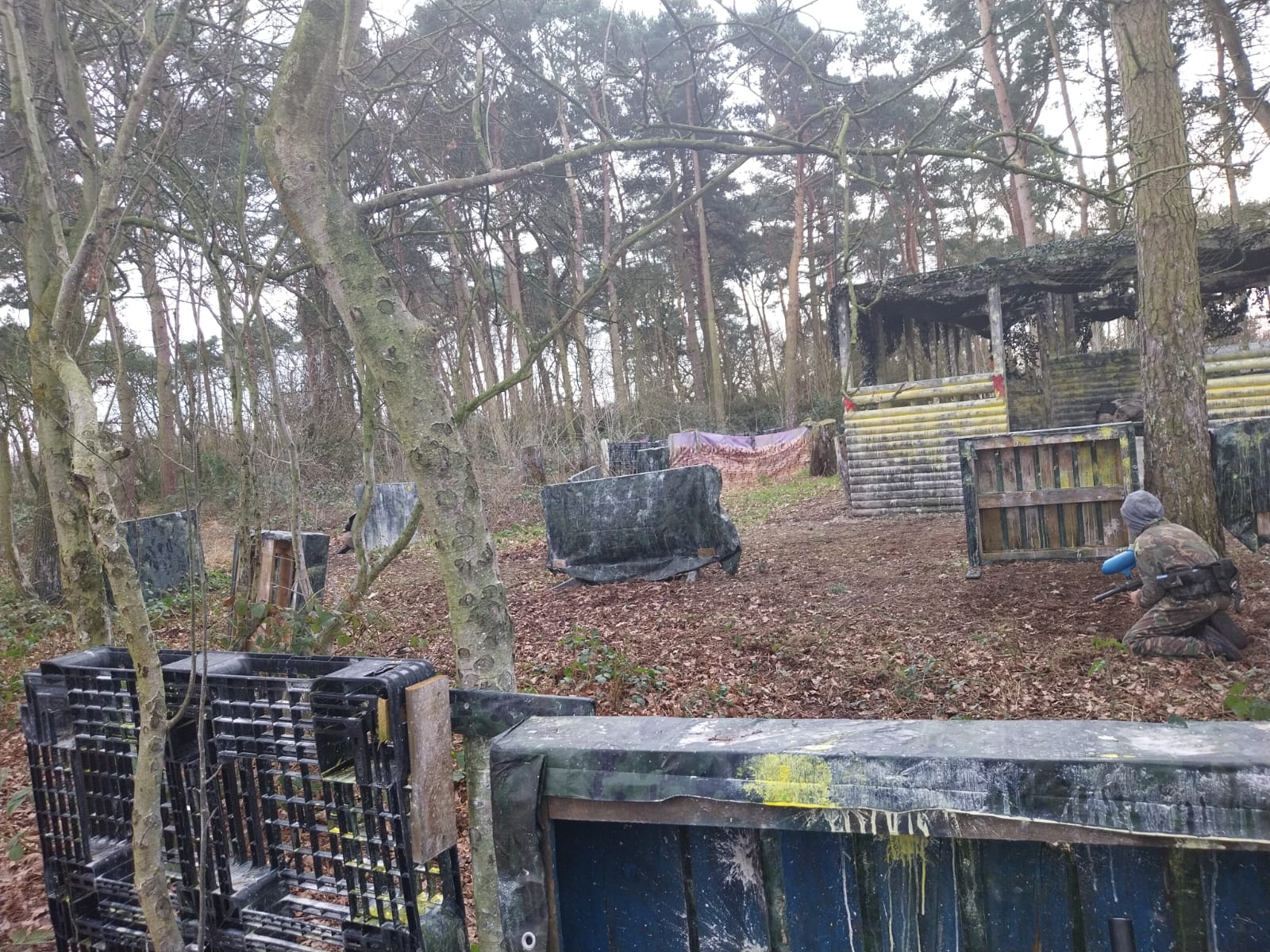 A person in advanced protective gear crouches behind a paint-splattered barrier in an adult version of a forested paintball arena, surrounded by trees and various makeshift obstacles, playing BlasterMasters' Mission Fortress Flag Heist.